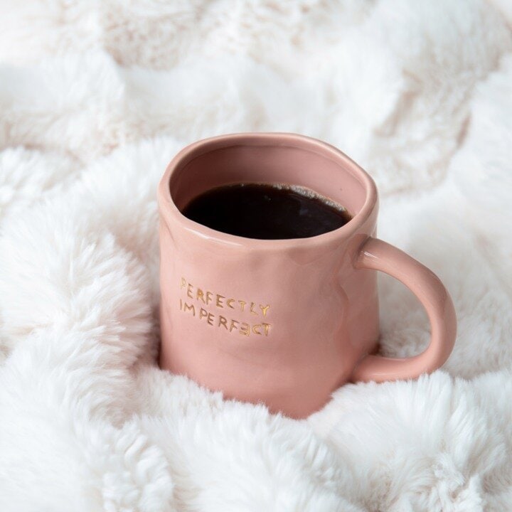 Rosa mugg med texten "Perfectly Imperfect" fylld med kaffe på en mjuk vit filt.
