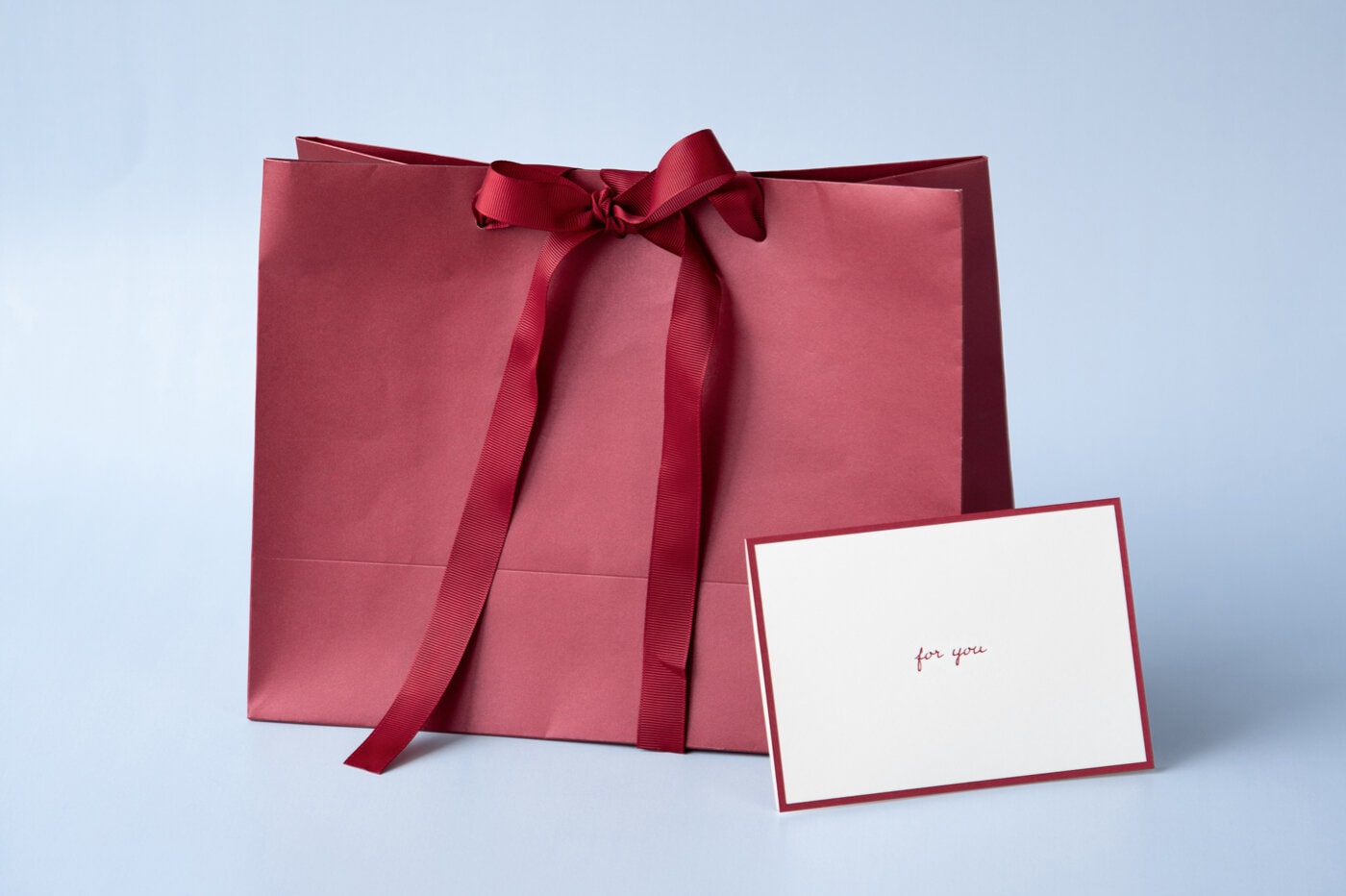 Vinröd presentpåse med rosett och presentkort framför ljus bakgrund.