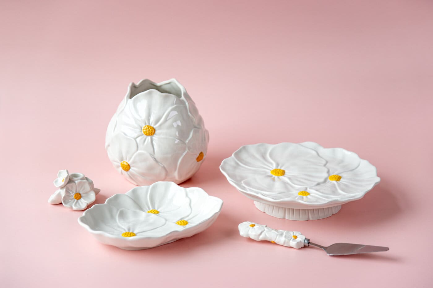 Vit keramikvas och serveringsfat med blomformade detaljer och gula mittpunkter ur serien Blomstra mot ljusrosa bakgrund.
