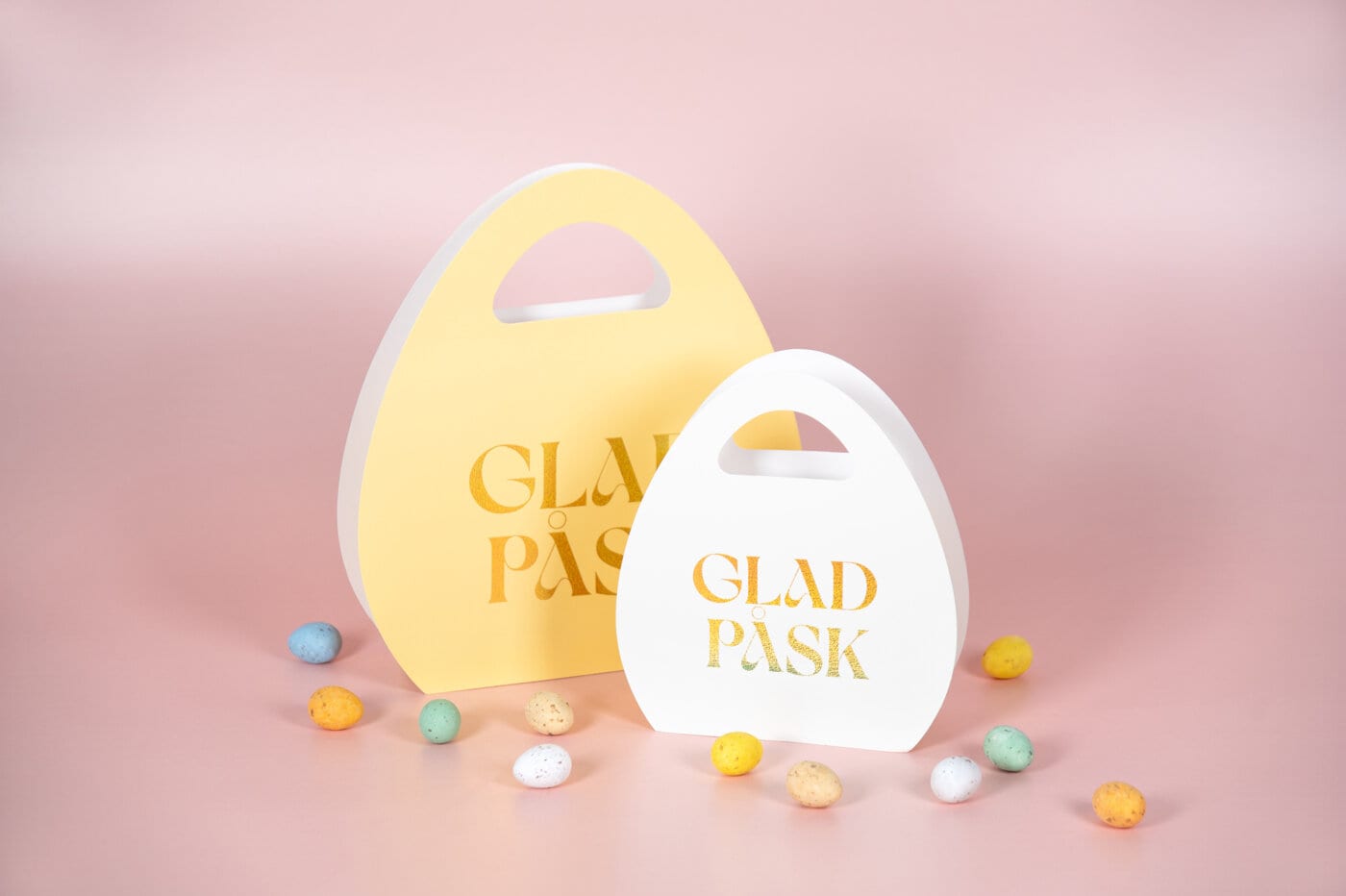 Äggformade påskpresentpåsar med texten ”Glad påsk”, omgivna av små färgade påskägg mot en mjuk rosa bakgrund.
