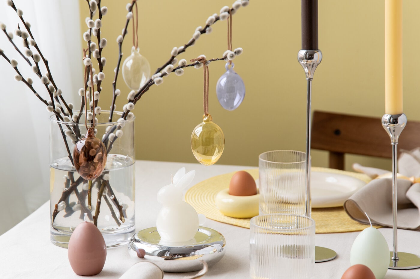 Påskdekorationer på bord med glasvas, videkvistar och hängande ägg i mjuka vårfärger