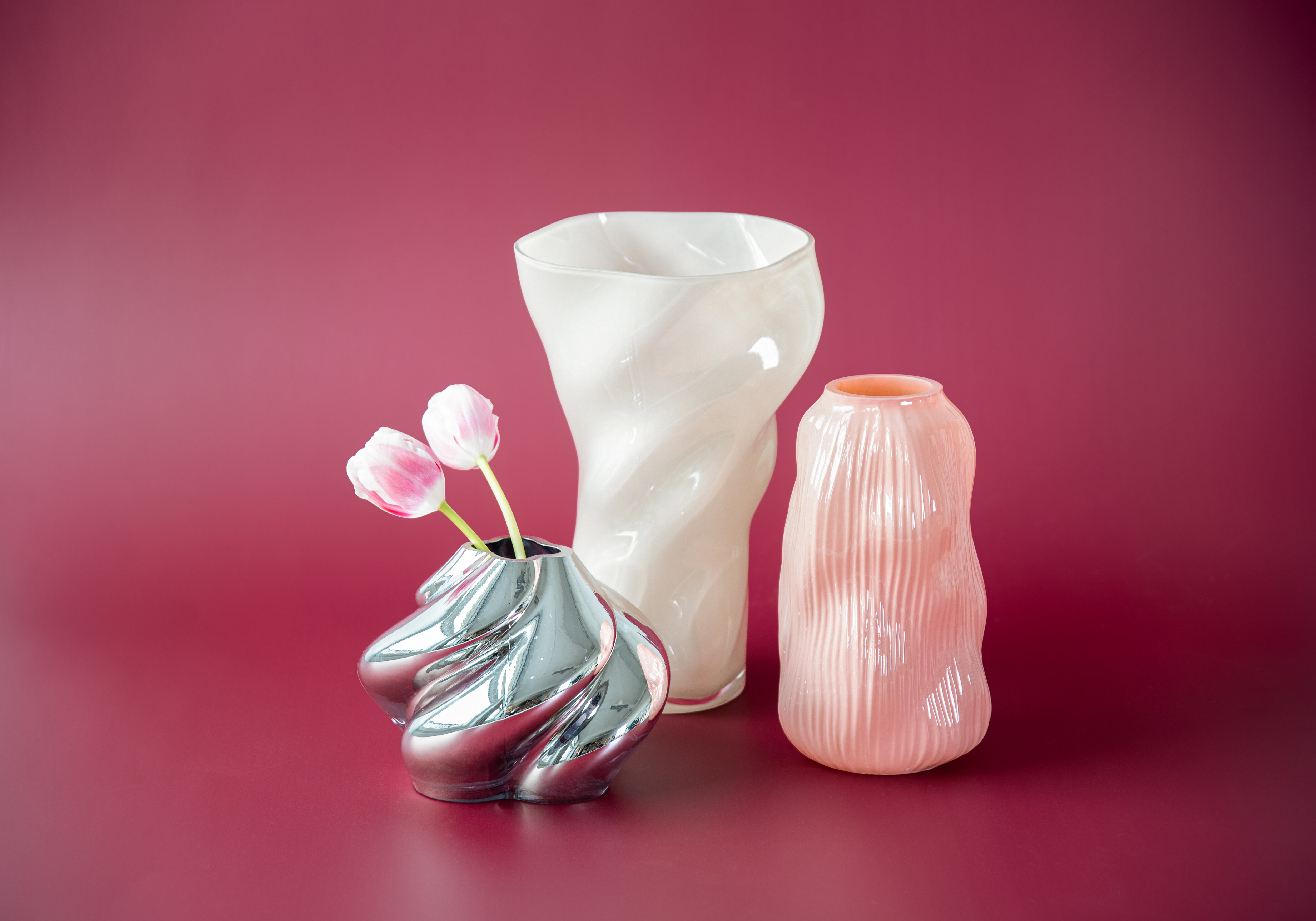 Tre vaser i olika färger och former mot vinröd bakgrund.