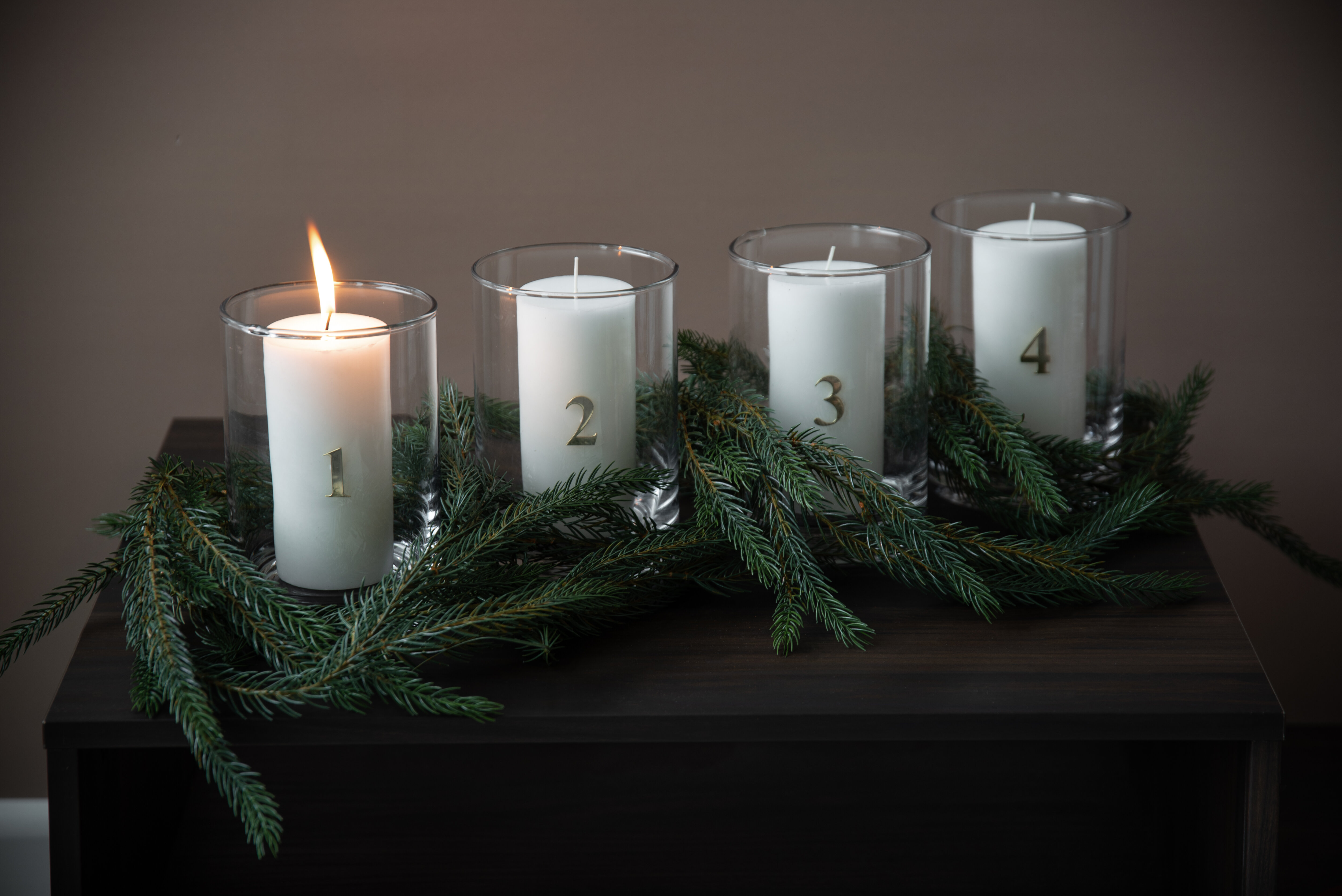 Fyra adventsljus i glaslyktor med siffror 1–4, dekorerade med grankvistar.