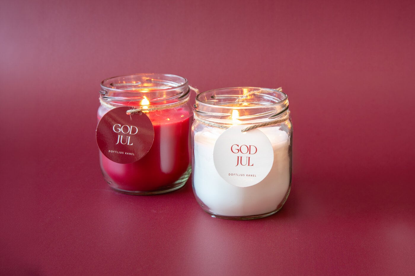 Två tända doftljus i glas, ett rött och ett vitt, med etiketten "God Jul" mot en röd bakgrund.