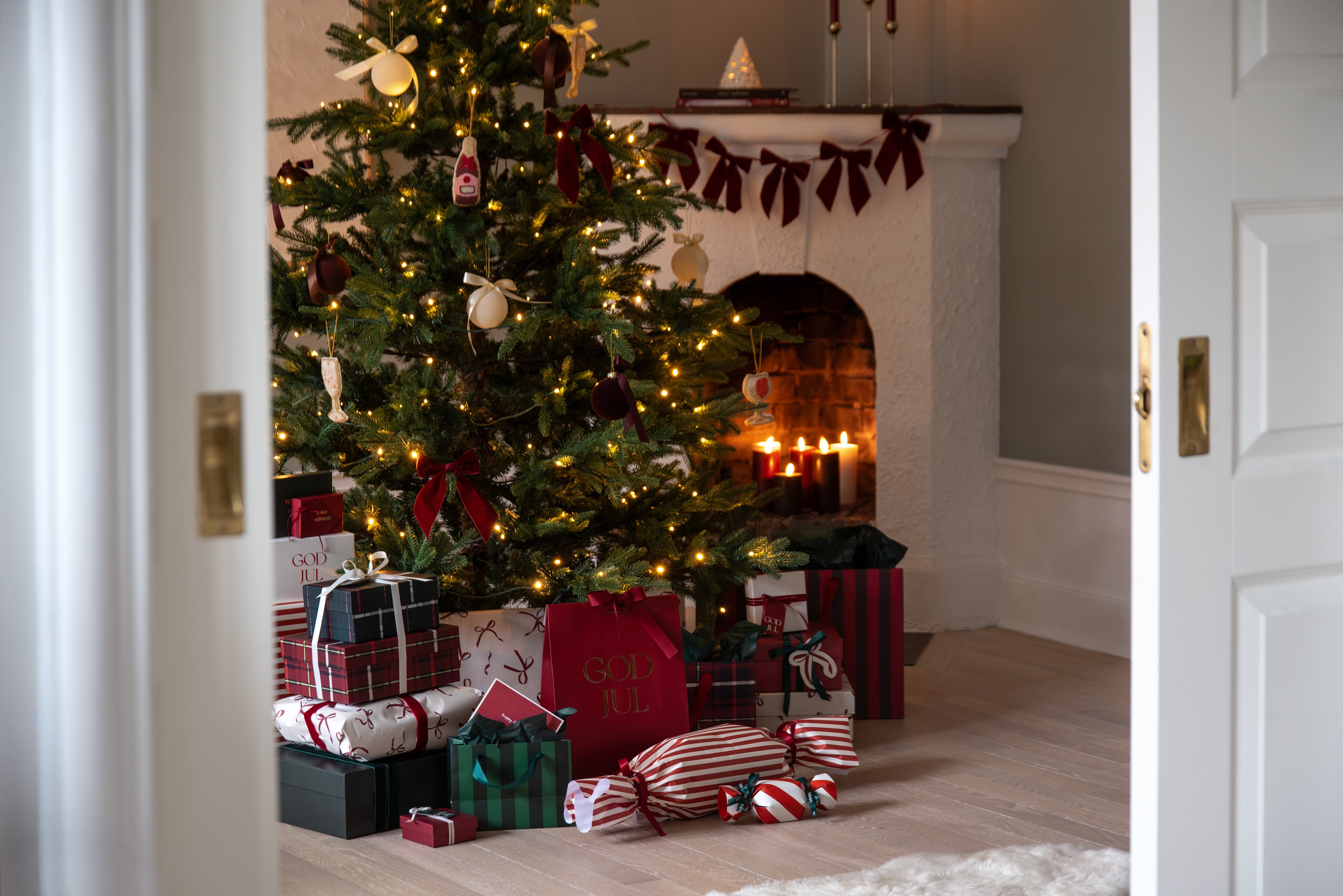 Klädd julgran med julklappar framför en öppen spis med tända ljus.