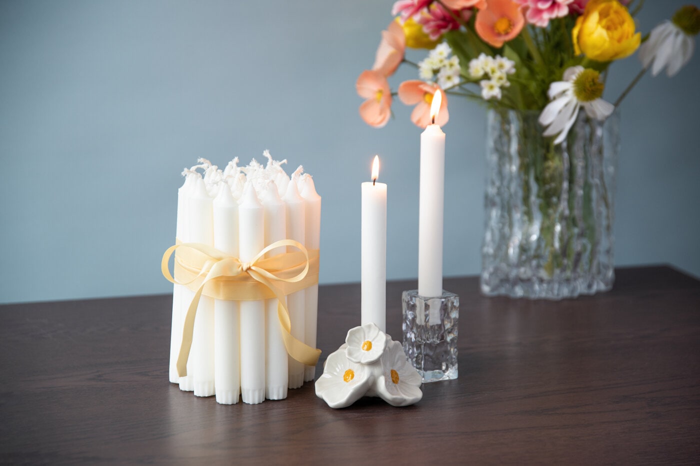 Bundna vita kronljus och tända ljus på ett bord med blommor i bakgrunden.