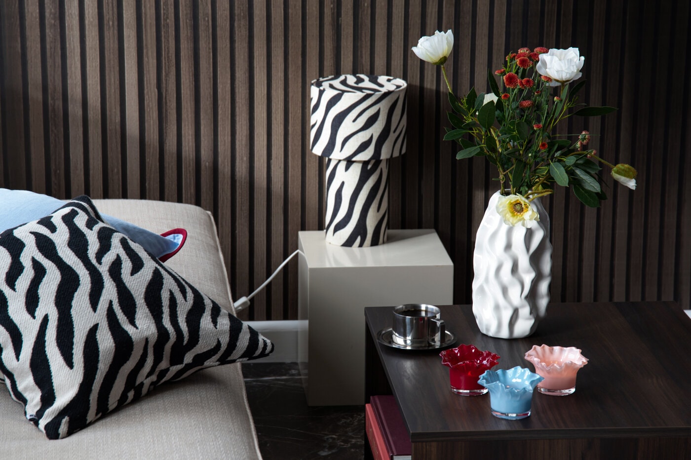 Inredningsmiljö med mönstrad bordslampa, mönstrad kudde och vit vas med blommor.