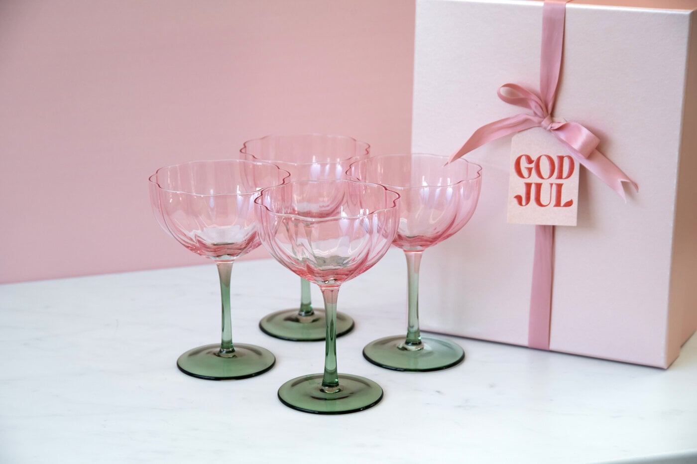Fyra coupeglas i rosa glas med gröna fötter uppställda på ett vitt bord bredvid ett paket inslaget i rosa band och en presenttag med texten “God jul”.