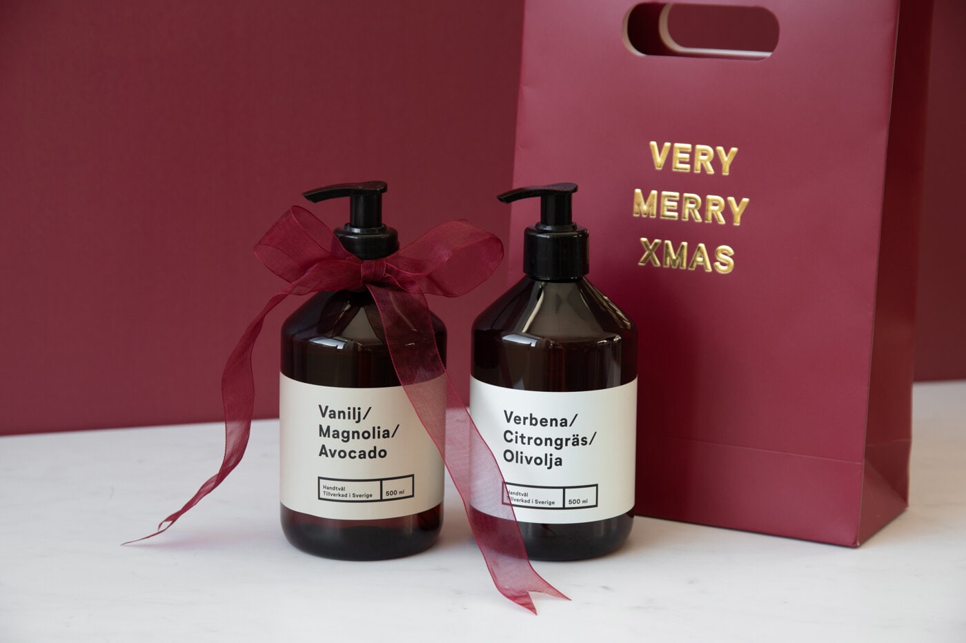 Två tvålflaskor, en dekorerad med rosett, bredvid en julpresentpåse.