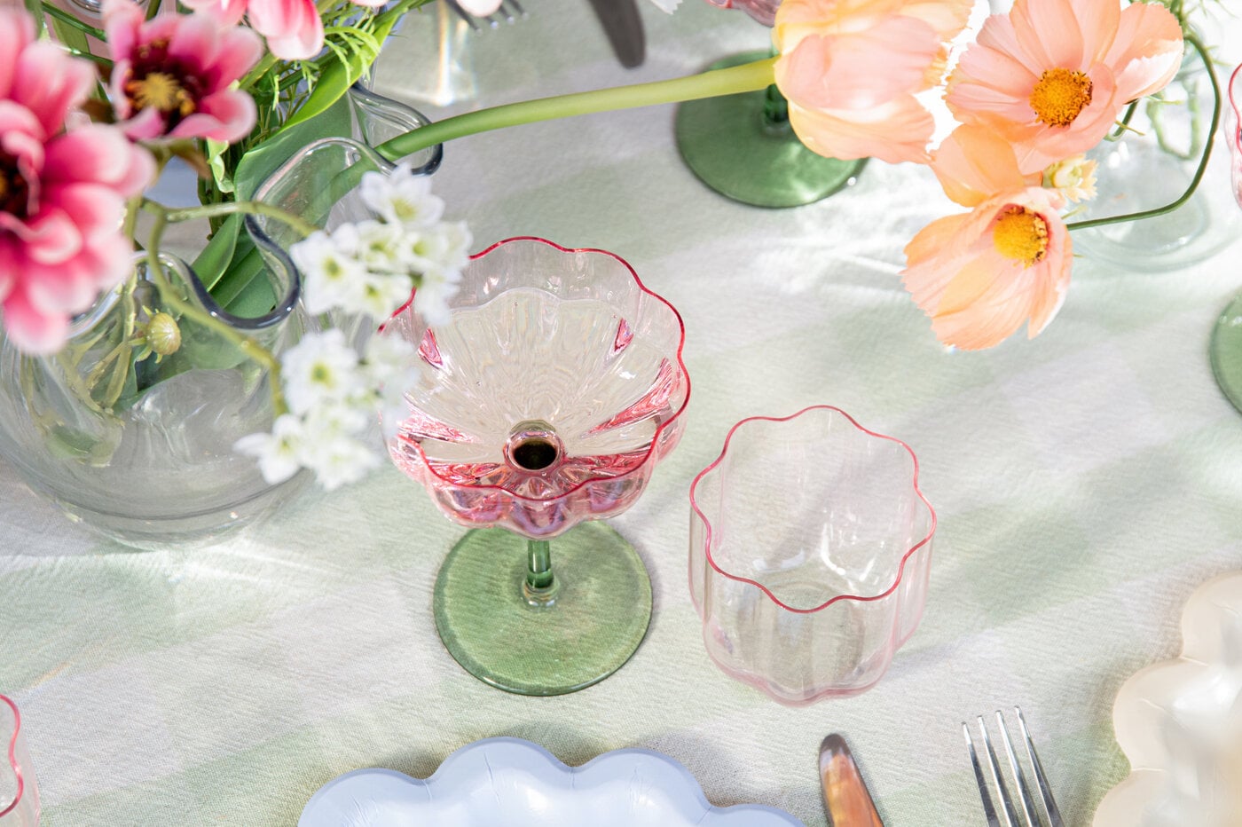 Dekorativa glas med vågig kant på dukat bord bland blommor.
