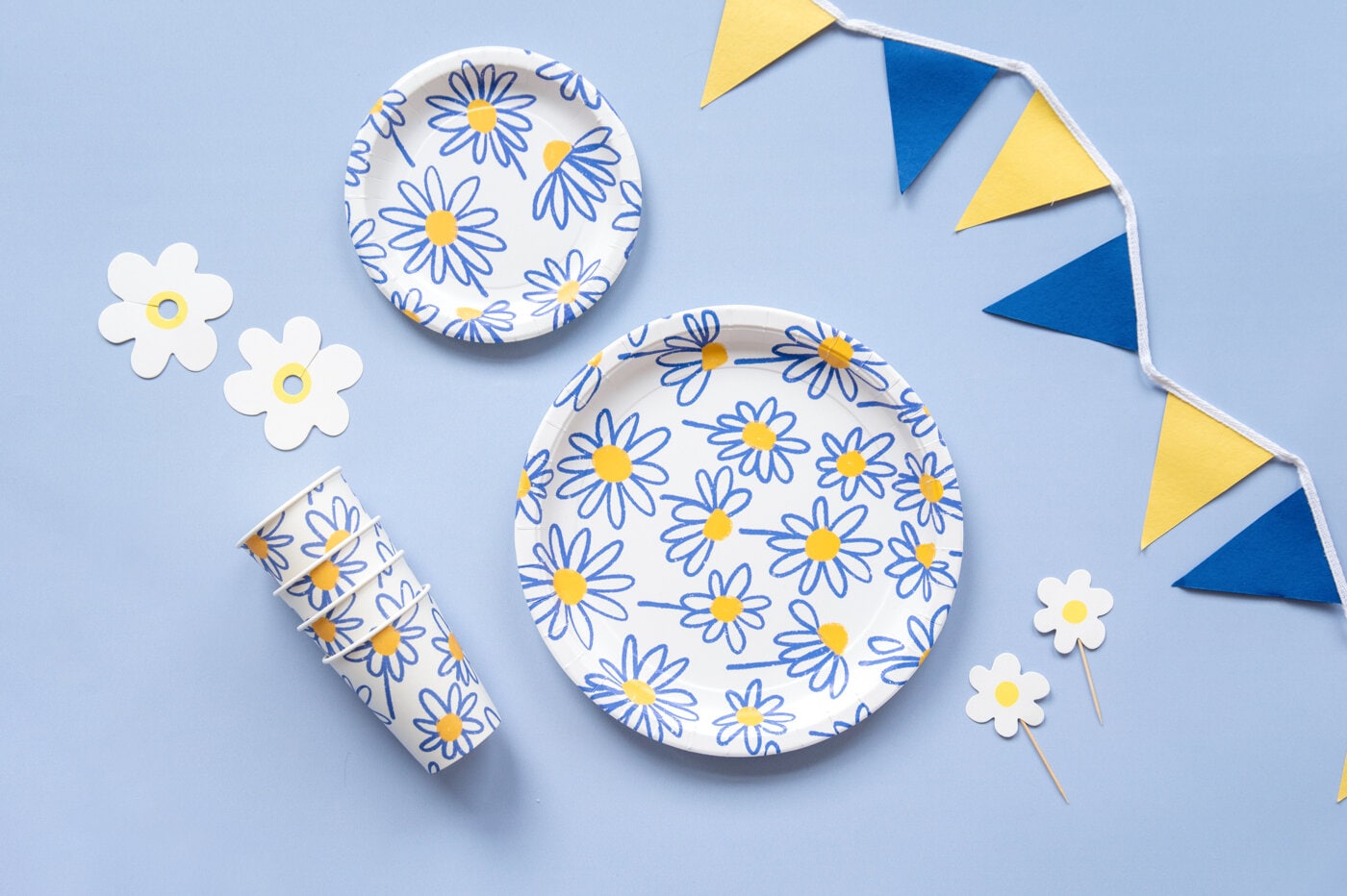 Blå‑gula pappersartiklar med prästkragemönster, inklusive tallrikar, muggar och små pappersdekorationer.