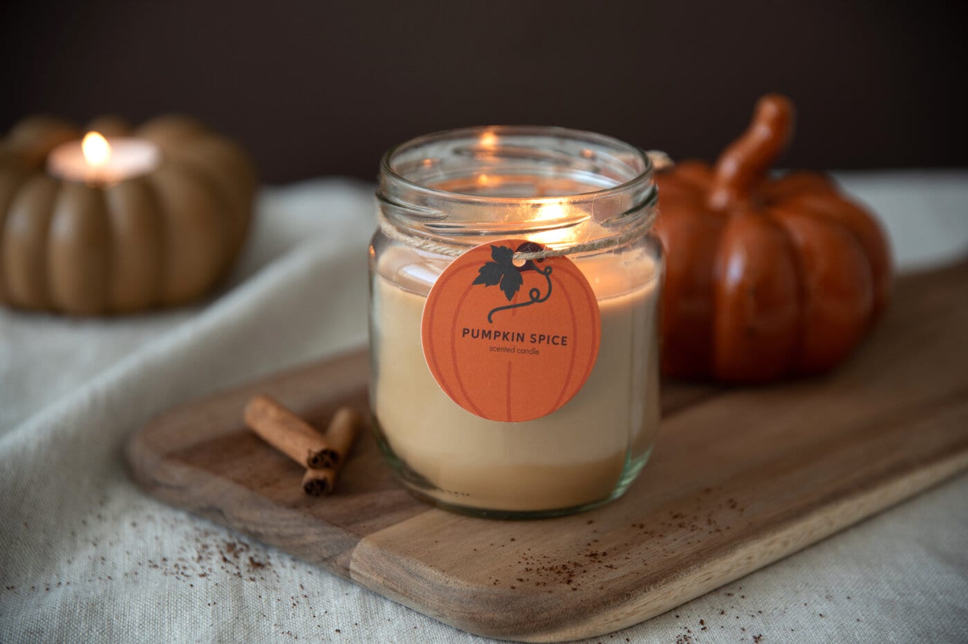Tänt Pumpkin Spice-doftljus i glas med pumpor och kanelstänger i bakgrunden.
