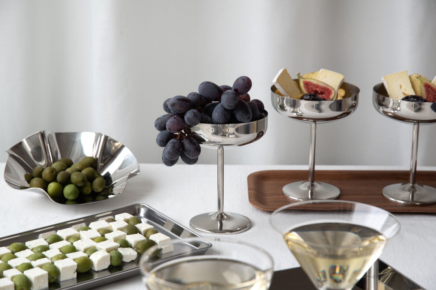 Silverfärgade coupeglas i metall med vindruvor, oliver, ost och fikon på ett festdukat bord.