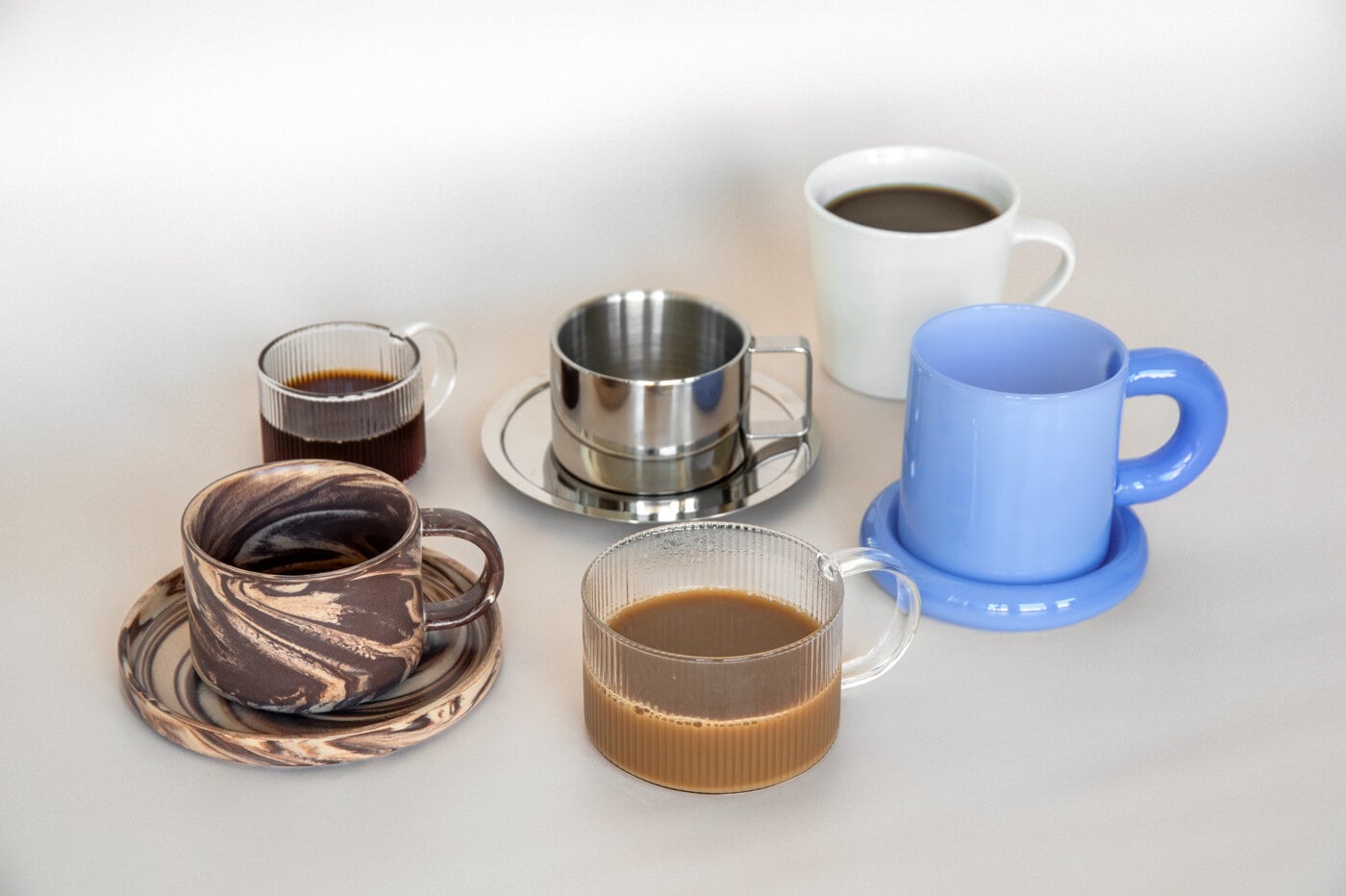 Muggar och koppar i glas, porslin och metall med kaffe, flera med matchande fat.