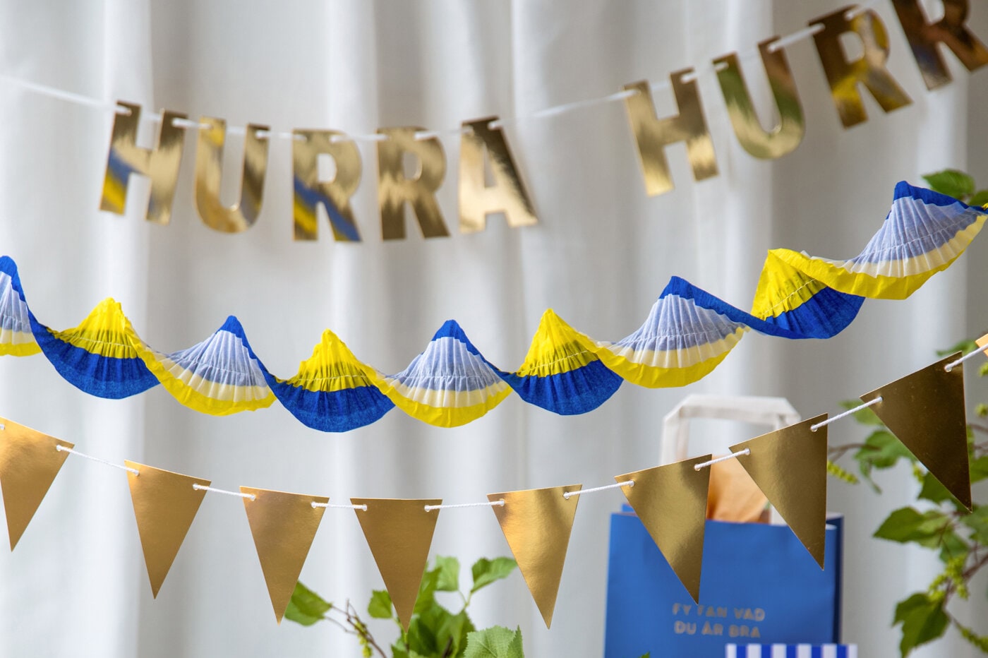 Blå‑gula studentdekorationer och guldfärgade vimplar och pappersgirlander över ett dukat bord inför studentfirande.